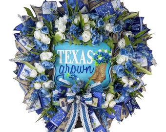Peut inclure: Une couronne ronde ornée de fleurs bleues et blanches, de rubans et de verdure. La couronne présente une enseigne centrale avec l'inscription "TEXAS grown" et une botte de cowboy. Un objet décoratif.