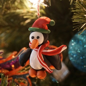 May include: A penguin Christmas ornament made of clay. The penguin is wearing a red and green Santa hat and a red and white scarf.