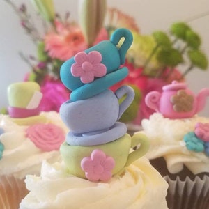 May include: Three cupcakes decorated with a teacup and teapot theme. The cupcakes are topped with white frosting and colorful fondant decorations. The cupcakes are on a white surface.