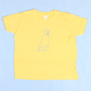 Op de afbeelding: Geel T-shirt met korte mouwen met een blauwe schets van een hond en de tekst "be brave" op de voorkant.