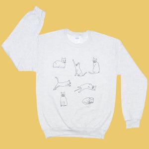 Op de afbeelding: Een grijs sweatshirt met een grafische print van acht katten in verschillende poses. De katten zijn zwart omlijnd en kijken allemaal naar voren.