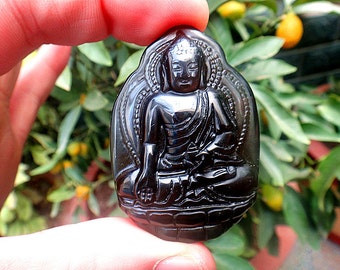 May include: Black obsidian pendant carved with a detailed image of a seated Buddha. The pendant is smooth and polished, with a glossy finish.