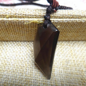 Op de afbeelding: Een zwarte obsidiaan hanger met een facetgeslepen ontwerp hangt aan een bruin koord. De hanger bevindt zich in een kleine doos met een bruine stoffen voering.