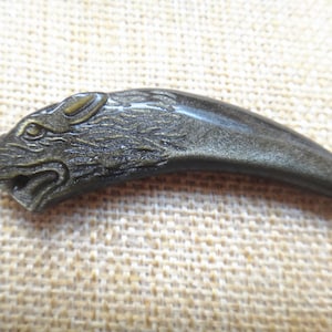 Op de afbeelding: Een gedetailleerde, metaalachtig uitziende klauw hanger met een wolfskop gebeeldhouwd aan de basis. De hanger is donker, gedempt goudkleurig en heeft ingewikkelde details. De klauw buigt naar een punt, en de wolfskop kijkt naar links.