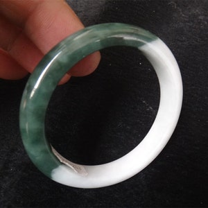 Può includere: Un braccialetto di giada verde e bianco. Il braccialetto è realizzato in pietra di giada liscia e lucida. La giada verde è di un verde più scuro da un lato e sfuma in un verde più chiaro dall'altro lato. L'altro lato del braccialetto è bianco.