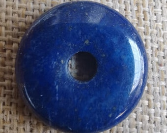 May include: A deep blue donut-shaped lapis lazuli stone with a hole in the center. The stone has a smooth, polished surface and is surrounded by a beige fabric background.