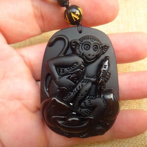 May include: Black obsidian pendant featuring a detailed carving of a monkey holding a peach. The pendant is attached to a black cord with gold beads.