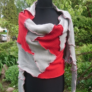 May include: A red and gray knitted wrap displayed on a black mannequin. The wrap features a patchwork design with wavy edges and a red interior. The sleeves are long and the background is a green garden.