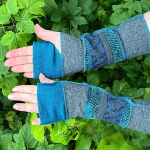 May include: Fingerless wrist warmers in shades of teal, gray, and blue. The warmers feature a textured design with a mix of knit and woven patterns. The background is a lush green foliage.