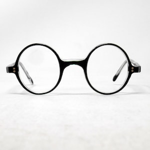 Op de afbeelding: Ronde bril met zwart montuur en heldere glazen. Het ontwerp is klassiek en minimalistisch. De bril staat tegen een effen witte achtergrond, waardoor de vorm wordt benadrukt. De glazen zijn rond.