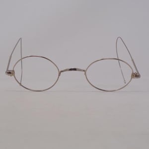 May include: A pair of silver-toned metal spectacles featuring thin, oval-shaped wire frames and long, slender, curved temple arms that extend outwards. The bridge is a simple, narrow metal arch connecting the two circular lens rims.