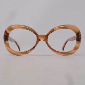 Può includere: Occhiali vintage oversize con un motivo marmorizzato marrone chiaro e marrone scuro. Le montature hanno una forma ovale arrotondata. Le aste e il ponte nasale sono di un marrone più scuro. Le lenti sono trasparenti.