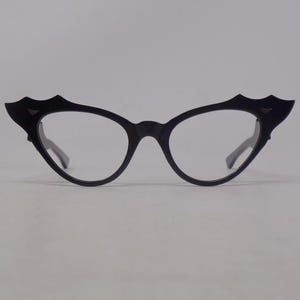 May include: Black cat-eye style eyeglasses with a unique bat-wing design on the upper frame. The glasses have clear lenses and a sleek, modern aesthetic. The frames are a matte black, offering a bold contrast against the clear lenses.