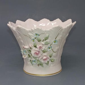 May include: A white porcelain bowl with a scalloped rim and a gold-trimmed base. The exterior features a raised relief design of light pink roses, green leaves, and small blue flowers arranged in a cluster on the front of the vessel.