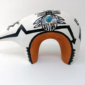 May include: A ceramic bear figurine featuring a white base with black geometric line art and a central circular motif. The underside of the bear is terracotta orange, and a small, round, blue turquoise-colored stone is embedded on its back.