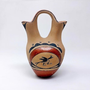 May include: A tan ceramic vessel with a bridge handle connecting two spouts. The body features hand-painted black and red geometric patterns, including a central circular motif containing a stylized black bird figure with wings extended.