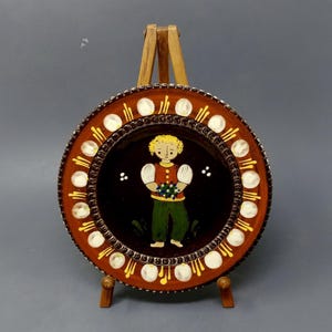 May include: A decorative plate with a brown rim and a black center, featuring a painted figure holding a bunch of blue grapes. The rim is adorned with white and yellow accents. The plate is displayed on a small wooden easel.