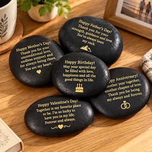 Puede incluir: Cinco piedras de río negras y lisas con texto e iconos grabados en color dorado. Cada piedra presenta un mensaje diferente para ocasiones como el Día de la Madre, el Día del Padre, cumpleaños, aniversarios y San Valentín, con símbolos.