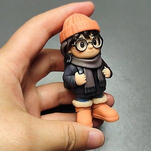 May include: A small figurine of a person wearing a dark blue coat, orange beanie, scarf, and glasses. The figure has a backpack and orange boots. The figurine is held in a hand against a gray background.