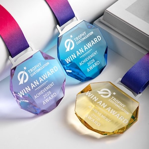 Puede incluir: Tres premios de cristal facetado con cintas coloridas. Cada premio tiene el texto "WIN AN AWARD" y "ACHIEVEMENT 2026 AWARD". Los premios son en tonos de azul, morado y dorado, con texto blanco y un soporte de cinta blanco.