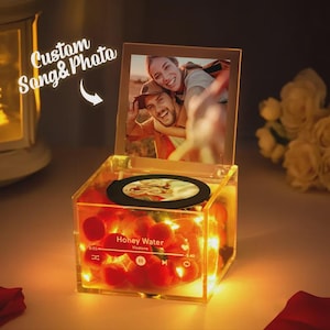 Könnte beinhalten: Eine transparente Acryl-Musikbox mit einem Foto und einer kleinen Disc. Die Box ist mit warmweißen Lichtern beleuchtet und mit roten Kugeln gefüllt. Der Text "Custom Song & Photo" ist sichtbar. Die Box hat ein Foto eines Paares.