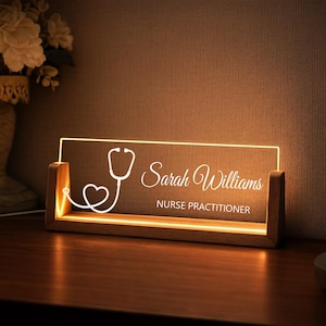 Puede incluir: Placa de escritorio personalizada con diseño de estetoscopio. El panel acrílico transparente está iluminado con un brillo cálido y presenta el nombre "Sarah Williams" y el título "Nurse Practitioner" en escritura blanca. La base es de madera.