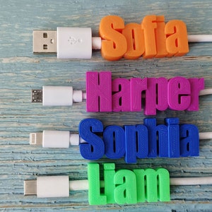 May include: Four personalized USB charging cables with names: Sofia (orange), Harper (pink), Sophia (blue), and Liam (green). Each name is 3D printed and attached to a white cable with a different USB connector type.