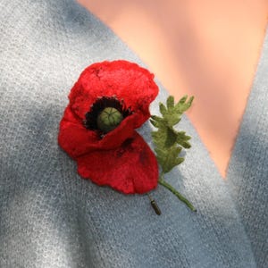 May include: A bright red poppy brooch with a dark centre and green detailing. The felt flower is pinned to a pale blue sweater. The brooch features a green stem and leaves.