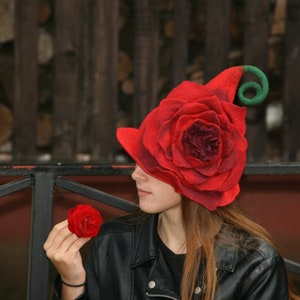 May include: A woman wearing a red felt rose hat with a green stem and a black leather jacket. She is holding a red felt rose in her hand.