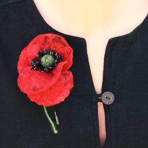 May include: A red felt poppy flower brooch with a dark green center and black stamens. The flower has a textured, layered appearance and is attached to a thin green stem with a silver-toned metal pin backing, secured to a black fabric garment.