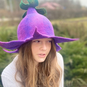 May include: A whimsical purple felt hat with a flower-like design. The hat features a wide brim and a central, bulbous crown with green accents. The hat is a unique accessory, perfect for costume or everyday wear.
