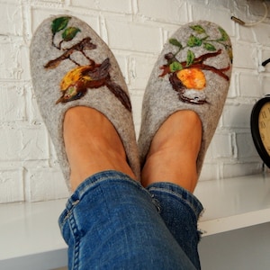 May include: A pair of gray felted wool slippers with a bird and branch design. The slippers feature a detailed hand-painted illustration of a bird perched on a branch with green leaves. The slippers are comfortable and cozy.