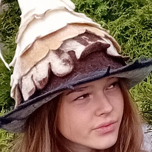 May include: A whimsical, layered felt hat in shades of white, tan, brown, and dark gray. The hat features a pointed crown and a wide brim, with a unique, textured design. The hat is set against a backdrop of green foliage.