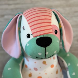 May include: A handmade plush dog toy with a pink and white striped head, green ears, and a white body with a pink and orange pattern. The toy has black button eyes and a black felt nose.