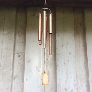 May include: A copper wind chime with five tubes hanging from a wooden ceiling. The chime has a small wooden tag hanging from the bottom.