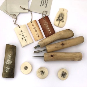 Può includere: Un set di strumenti per intagliare in osso e legno con disegni intricati. Gli strumenti includono un piccolo coltello, uno scalpello e un raschietto. I pezzi di osso sono decorati con motivi geometrici in inchiostro nero. Gli strumenti sono disposti su una superficie bianca.