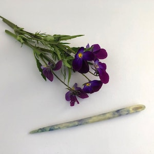 Op de afbeelding: Een takje paarse viooltjes met gele centra en groene bladeren naast een lichtblauwe en gele houten breinaald. De naald is aan één uiteinde puntig en heeft een afgerond uiteinde.