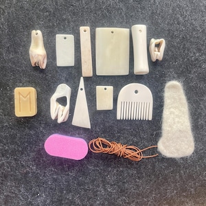May include: A collection of craft supplies including various off-white bone-colored pendants, a small comb, a rectangular block with an engraved M, a pink rectangular sanding block, a coil of brown leather cord, and a white felted wool piece.