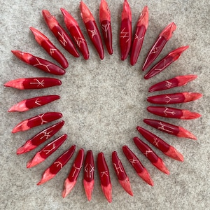 Może przedstawiać: Zestaw 24 czerwonych drewnianych run z białymi znakami. Runy są ułożone w kółko na szarym tle.