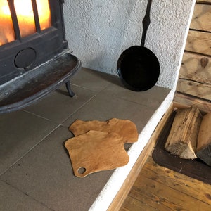 Op de afbeelding: Een gezellige scène met een open haard met vuur. Twee bruine leren pads met een gat liggen op de vloer. Een zwarte pan hangt aan de muur. Brandhout is in de buurt opgestapeld, wat wijst op een warme, rustieke omgeving.