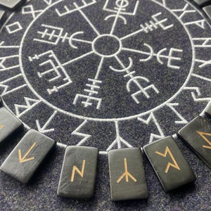May include: A set of black rune stones with gold symbols arranged around a circular, white, geometric design on a dark blue surface. The runes are rectangular and appear to be made of stone or a similar material.
