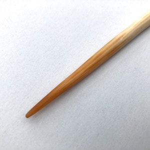 Könnte beinhalten: Ein Haarstab aus Holz mit konischem Design. Der Stab ist hellbraun mit dunkleren braunen Maserungsmustern. Die Spitze ist ein dunklerer Braunton. Der Hintergrund ist eine schlichte weiße Oberfläche.