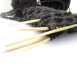 May include: Close-up of knitting in progress. Dark grey yarn is held on light-coloured wooden knitting needles. The needles are inserted into the knitted fabric, with some needles extending out. The background is white.