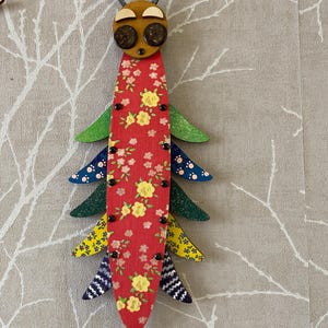 Könnte beinhalten: Ein farbenfrohes Holz-Insekt mit rotem Körper, grünen, blauen, gelben und weißen Flügeln und einem braunen Kopf mit schwarzen Augen. Das Insekt ist mit einem Blumenmuster verziert.