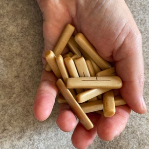 Op de afbeelding: Een hand die een stapel kleine, lichtbruine houten stokjes vasthoudt met een enkele lijn gegraveerd over elk stokje.
