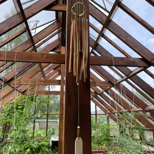 Op de afbeelding: Een handgemaakte windgong van natuurlijk hout hangt in een houten kas. De windgong heeft meerdere houten staven die aan een cirkelvormig frame zijn opgehangen, met een lang, rechthoekig houten stuk aan de onderkant. De kas heeft glazen panelen en houten balken.