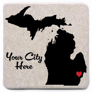 May include: Square stone coaster featuring a black silhouette of the state of Michigan. The text "Your City Here" is written in a cursive font. A red heart marks a location on the lower peninsula. The coaster has a light beige, speckled background.