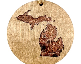 May include: A round wooden ornament with a laser-etched outline of the state of Michigan. The etching is a brown color and the wood is a light brown color.