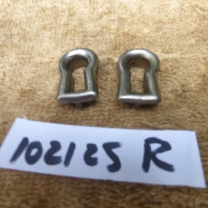 Könnte beinhalten: Zwei kleine, silberfarbene Schlüssellochschilder. Die Schilder sind aus Metall und haben die klassische Schlüssellochform. Unter den Schildern befindet sich ein weißes Etikett mit dem Text "102125 R".