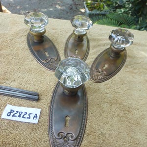 May include: Four antique door knobs. Each has a clear, faceted glass knob and an ornate, bronze-coloured backplate. The backplates feature a keyhole and decorative detailing. A small metal piece and a tag with the text "82825 A" are also visible.